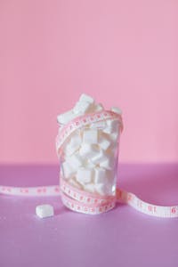 A glass of sugar cubes wrapped in measuring tape on a pink background symbolizes health awareness.