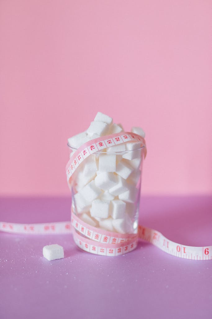 A glass of sugar cubes wrapped in measuring tape on a pink background symbolizes health awareness.