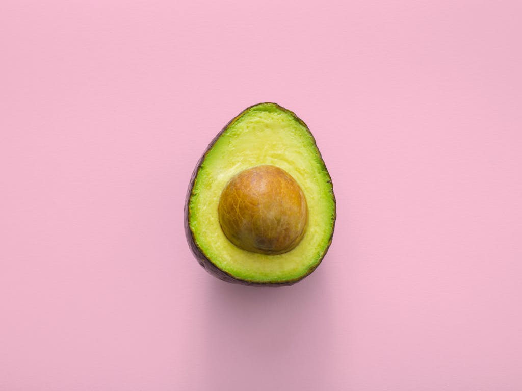 Fresh avocado fruit cut in half, showcasing seed, on a pastel pink background.